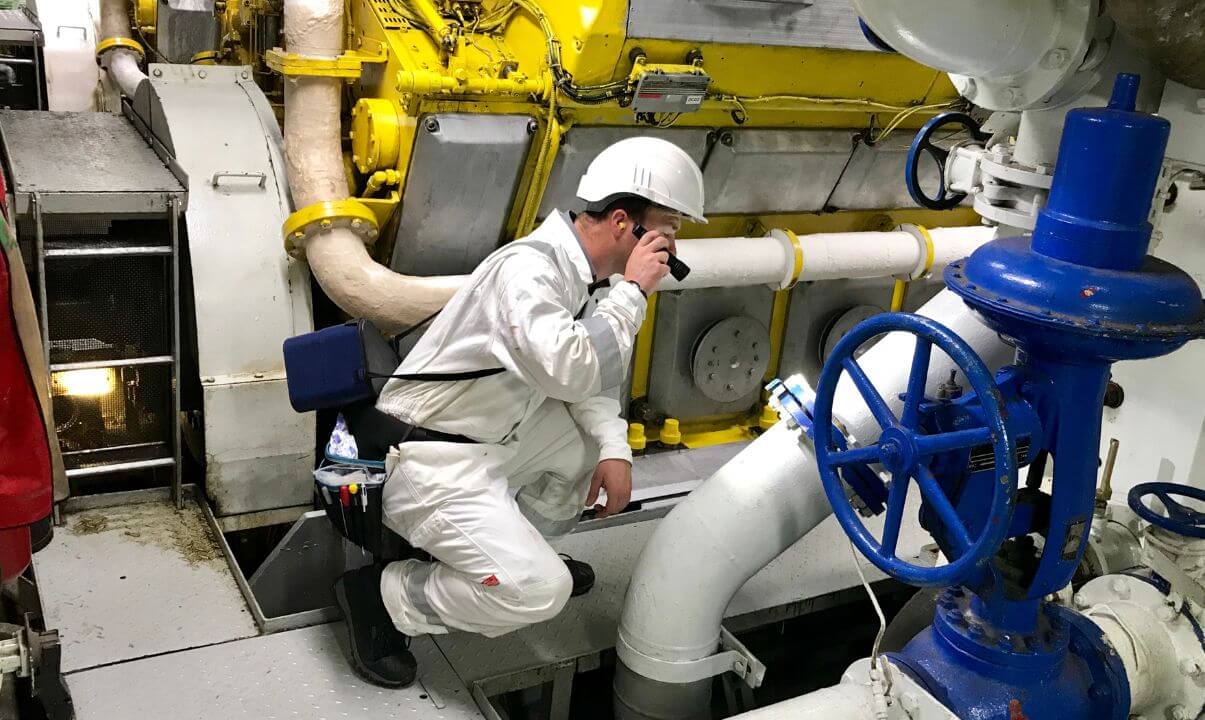 A man dressed in PPE full white body suit and helmet crouched down shining a torch on white piping in an enclosed engine room.